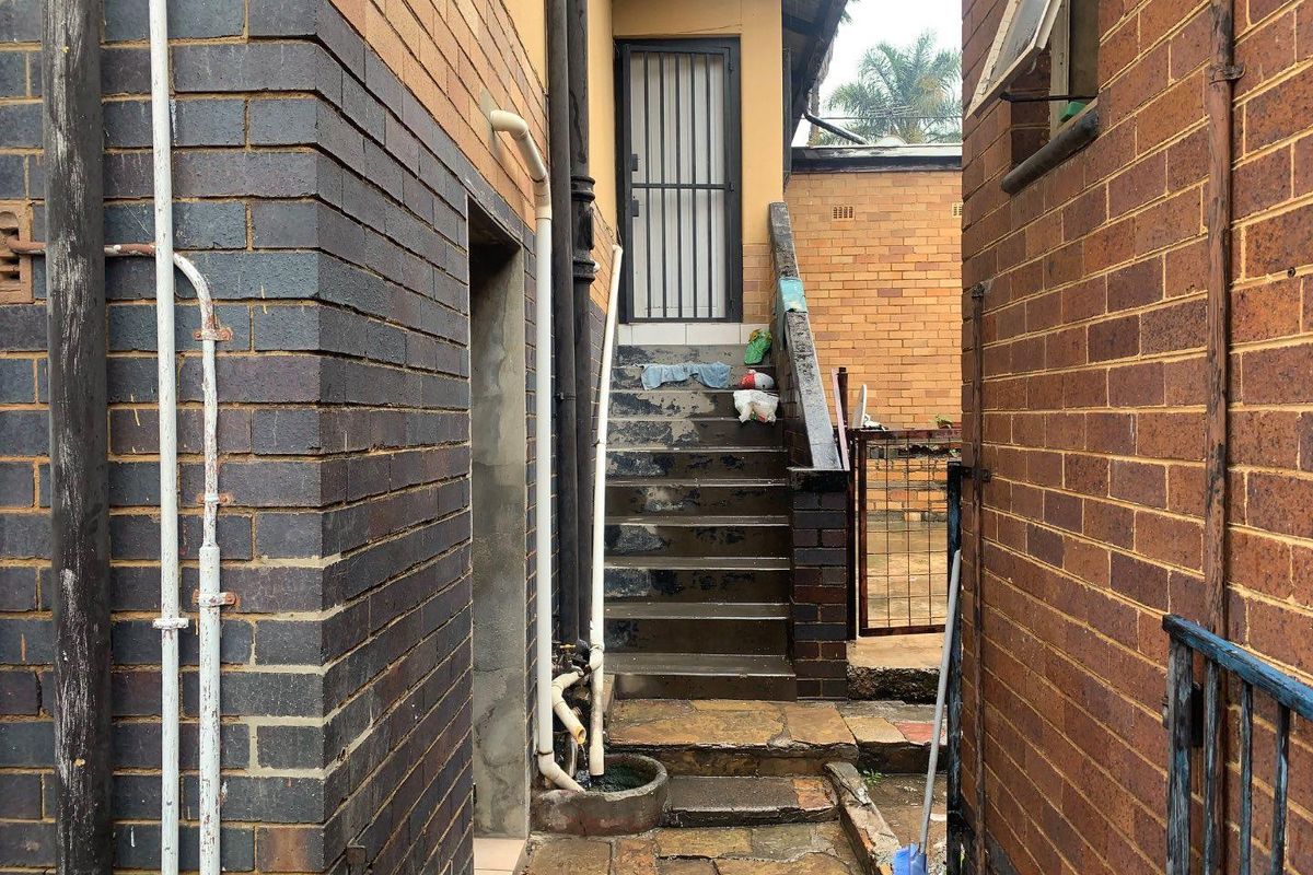 A narrow exterior side passage featuring brick walls, concrete stairs leading to a security door, and exposed utility piping.