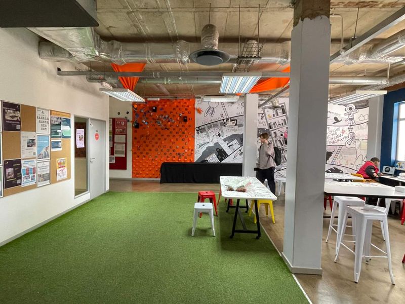 Open-plan office space featuring exposed industrial ceilings, ductwork, structural columns, and artificial turf flooring, creating a modern collaborative work environment.