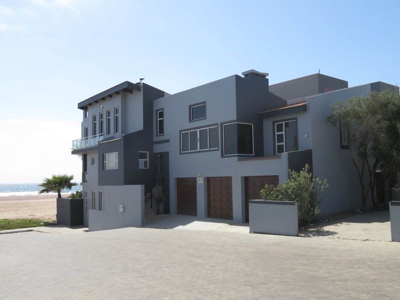 A modern multi-level beachfront house featuring a triple garage, paved driveway, and a balcony with glass railings overlooking the ocean.