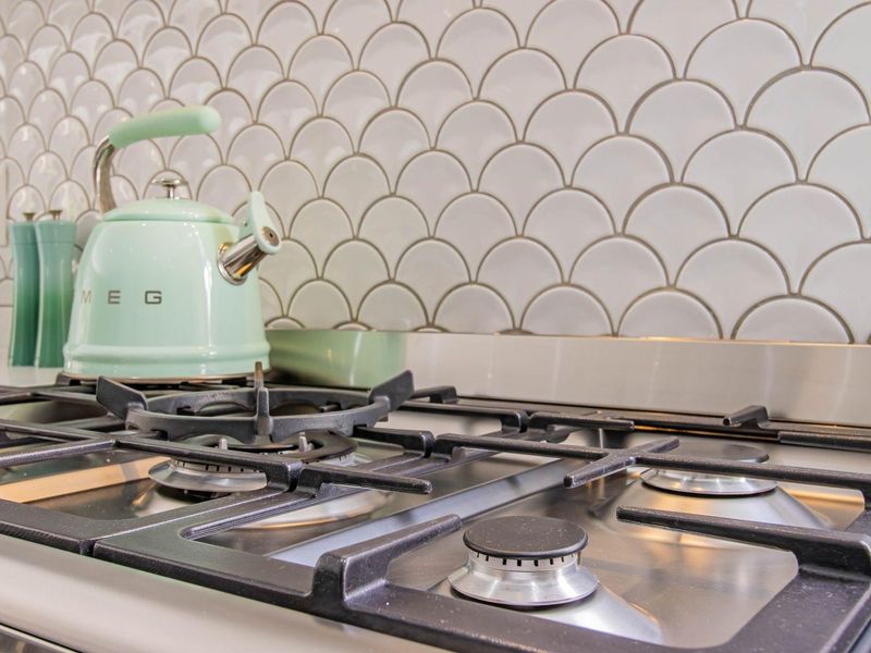 Close-up view of a modern kitchen featuring a stainless steel gas hob and a decorative white scalloped tile backsplash.