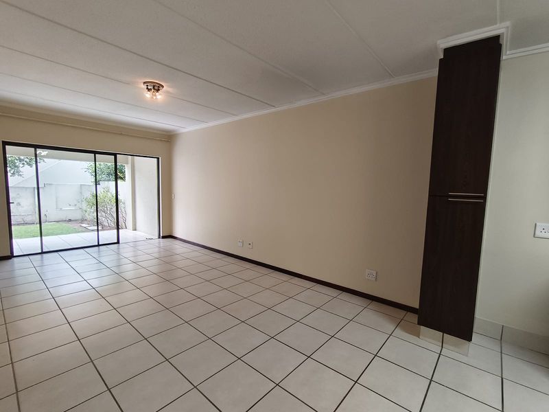 A spacious, empty living area featuring neutral tiled flooring, a built-in dark wood cupboard, and large sliding glass doors leading to an outdoor patio.