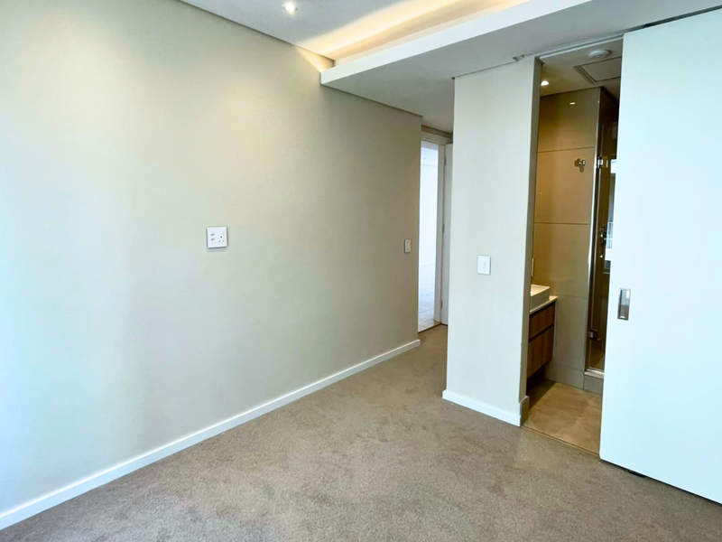 A clean, empty bedroom featuring neutral carpeting, recessed ceiling lighting, and a sliding door leading to an en-suite bathroom.