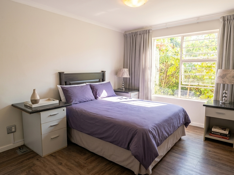 A well-maintained bedroom featuring laminate flooring, neutral walls, and a large window with security bars, providing natural light and a pleasant garden view.