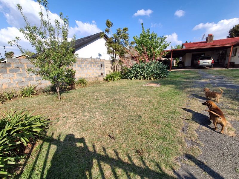 A spacious residential garden featuring a lawn area, concrete block boundary wall, and an adjacent covered carport with a driveway.