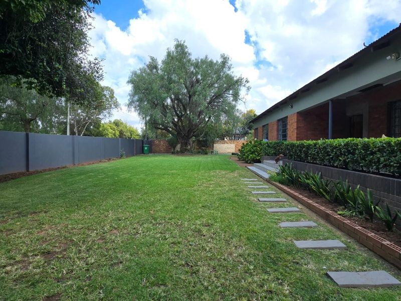 A well-maintained rear garden featuring a spacious lawn, perimeter wall, and a paved pathway leading along the brick-faced house.