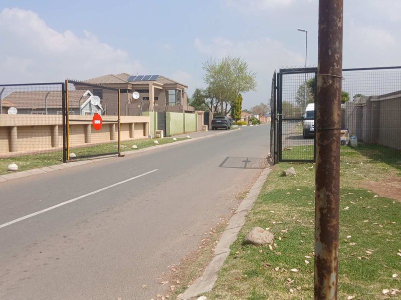 A street view showing the entrance to a residential area with perimeter walls, security gates, and solar panels visible on nearby rooftops.