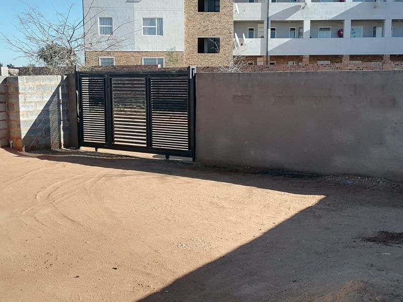 A rear exterior view showing an unpaved yard area enclosed by a perimeter wall and a modern black metal sliding gate.