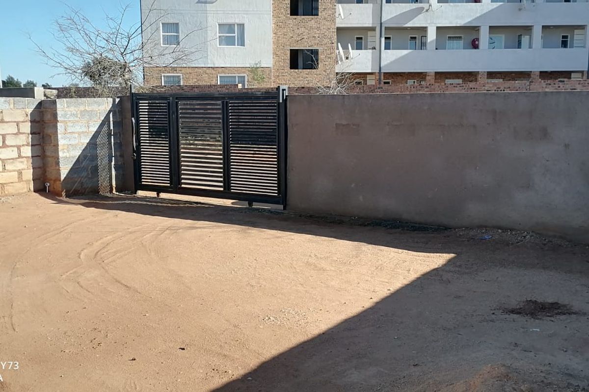 A rear exterior view showing an unpaved yard area enclosed by a perimeter wall and a modern black metal sliding gate.