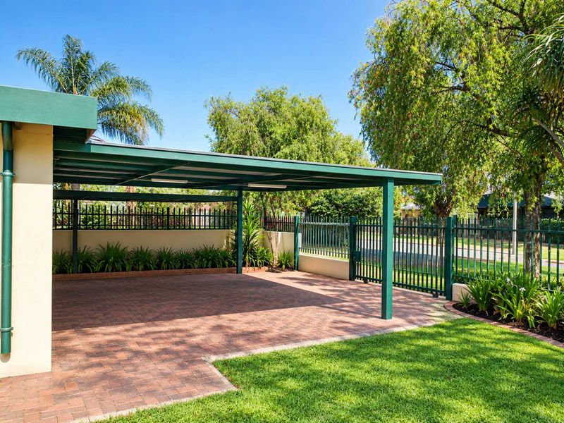 A well-maintained paved carport area featuring a metal roof structure, secure perimeter fencing, and established garden landscaping.