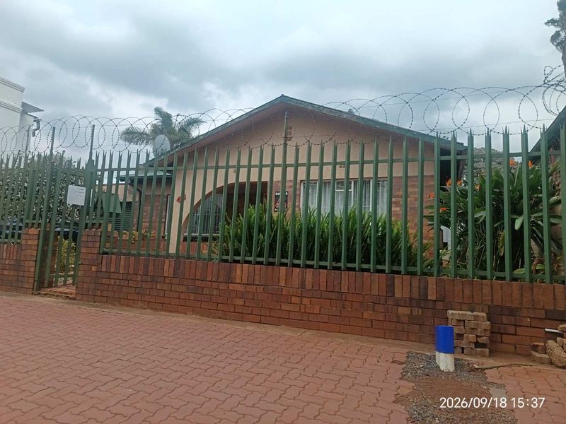 Front exterior view of a brick house featuring a secure perimeter with steel palisade fencing, razor wire, and a paved driveway.