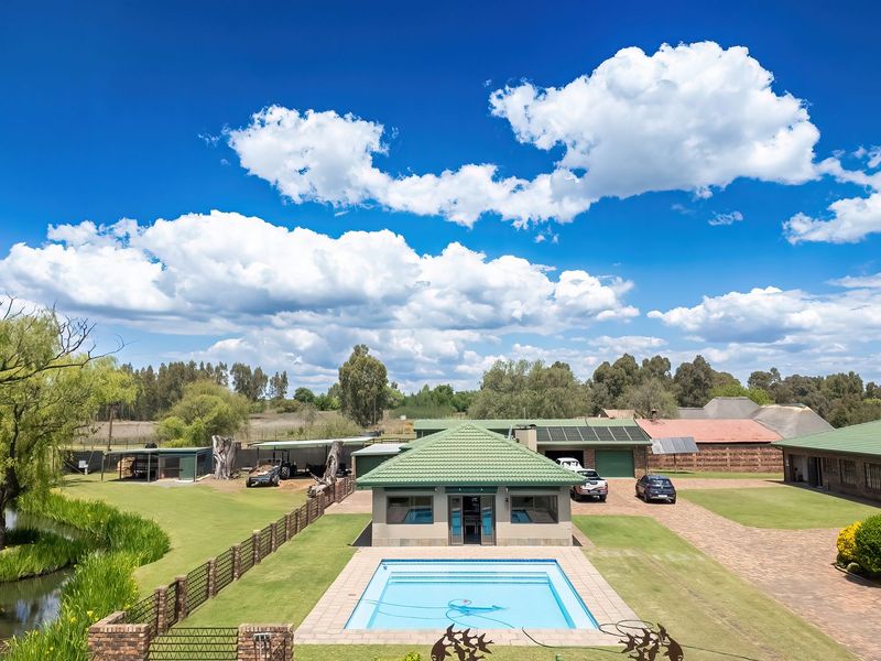 Aerial view of a spacious property featuring a swimming pool, extensive paved driveway, landscaped gardens, a pond, and multiple outbuildings with green tiled roofs.