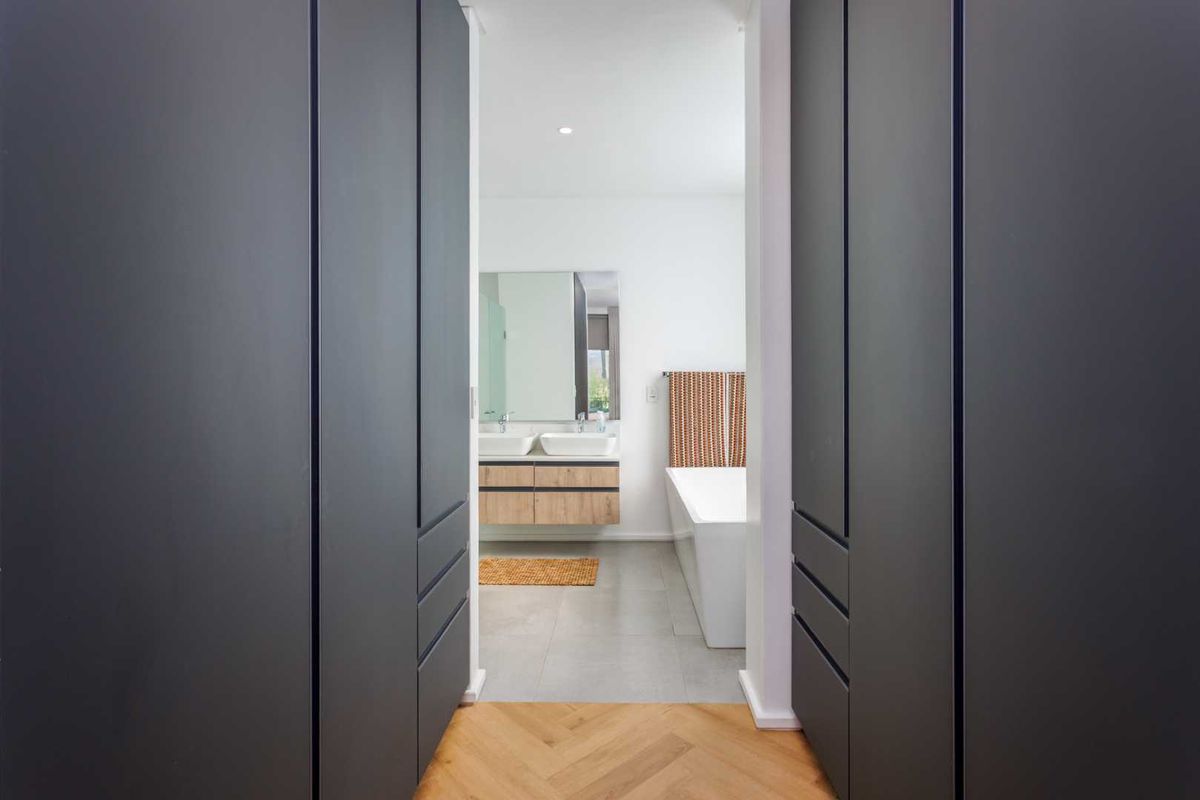 A modern bathroom featuring a double vanity with wood-finish cabinetry, a freestanding bathtub, and large-format floor tiles, viewed from an adjacent hallway.