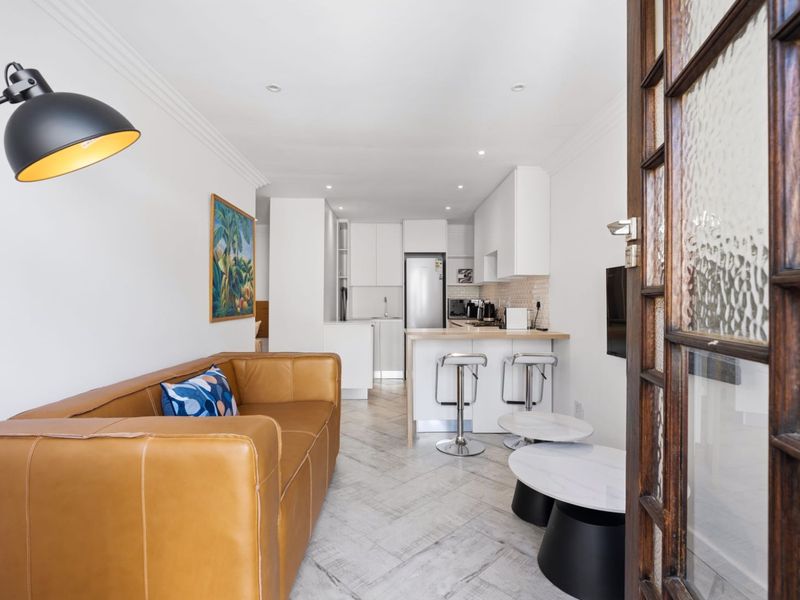 A bright open-plan living and kitchen area featuring modern cabinetry, a breakfast island, and tiled flooring, viewed through a classic timber door frame.