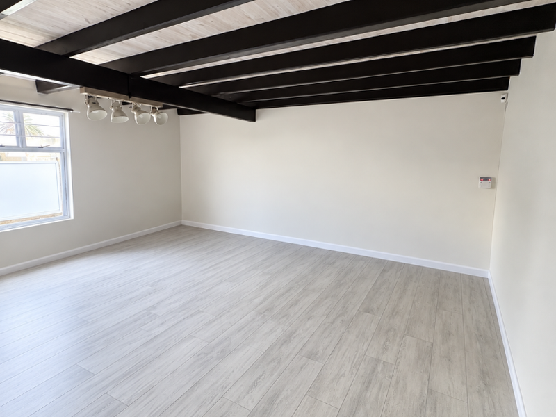 A bright, empty living room featuring light wood-look laminate flooring and striking exposed dark timber ceiling beams with integrated track lighting.