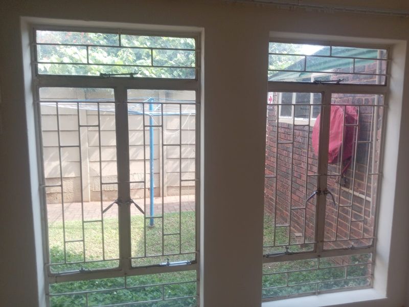 Interior view of a bedroom featuring steel-framed windows with security bars, overlooking a rear garden area.