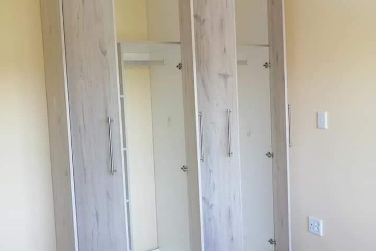 A bright bedroom featuring modern light wood-finish built-in wardrobes and neutral tiled flooring.