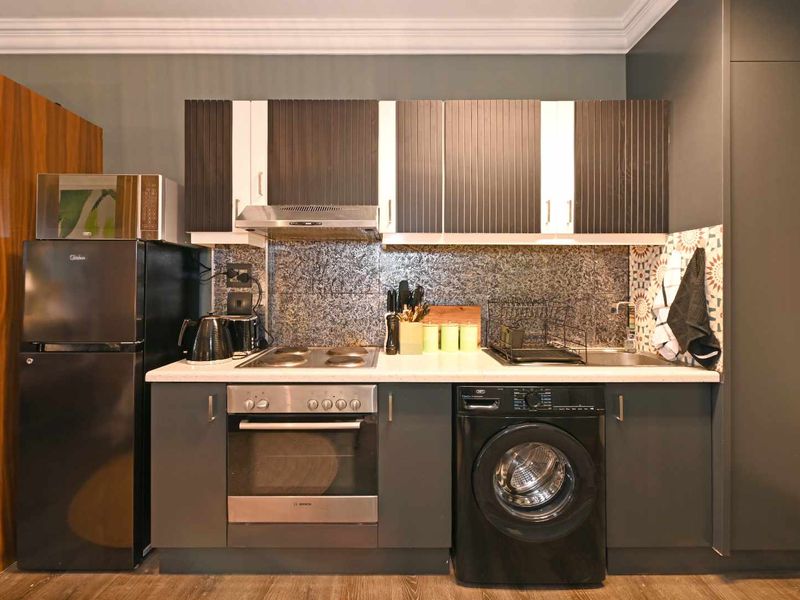 Compact modern kitchen featuring dark cabinetry, stone countertops, built-in oven, electric hob, extractor fan, and an integrated washing machine.