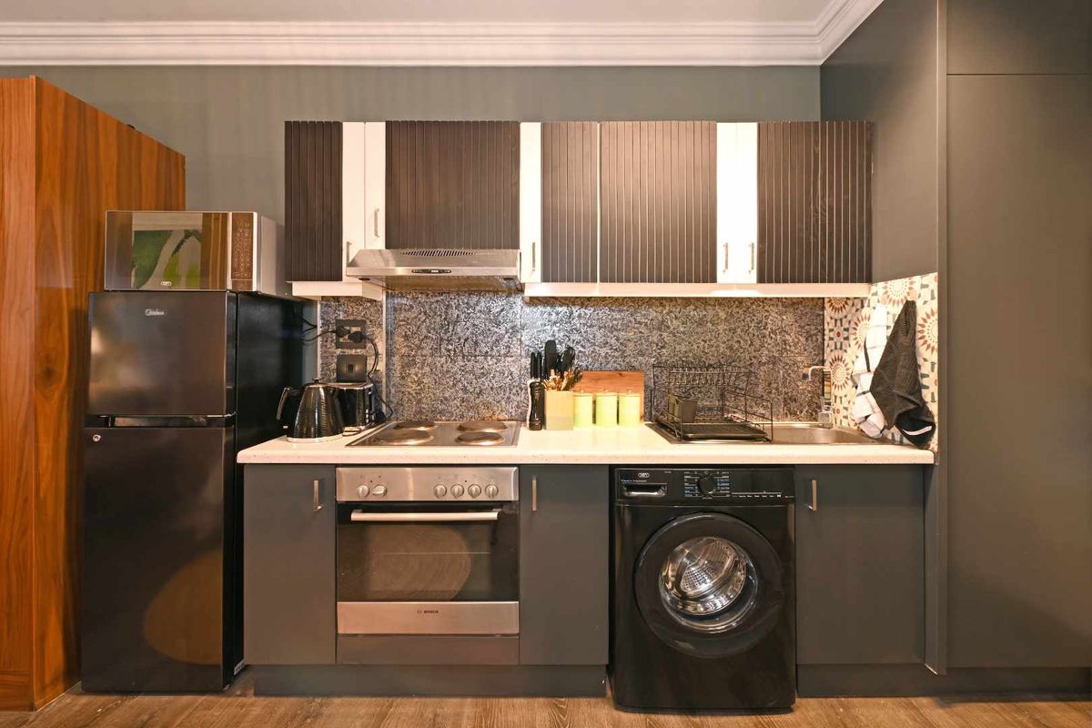 Compact modern kitchen featuring dark cabinetry, stone countertops, built-in oven, electric hob, extractor fan, and an integrated washing machine.