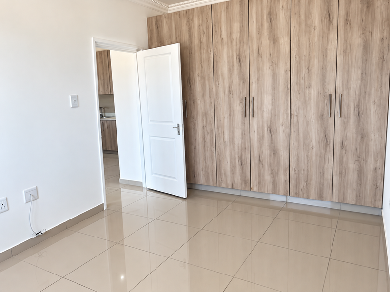 A bright, empty bedroom featuring modern wood-finish built-in cupboards, polished floor tiles, and clean white walls, offering a neutral space ready for furnishing.