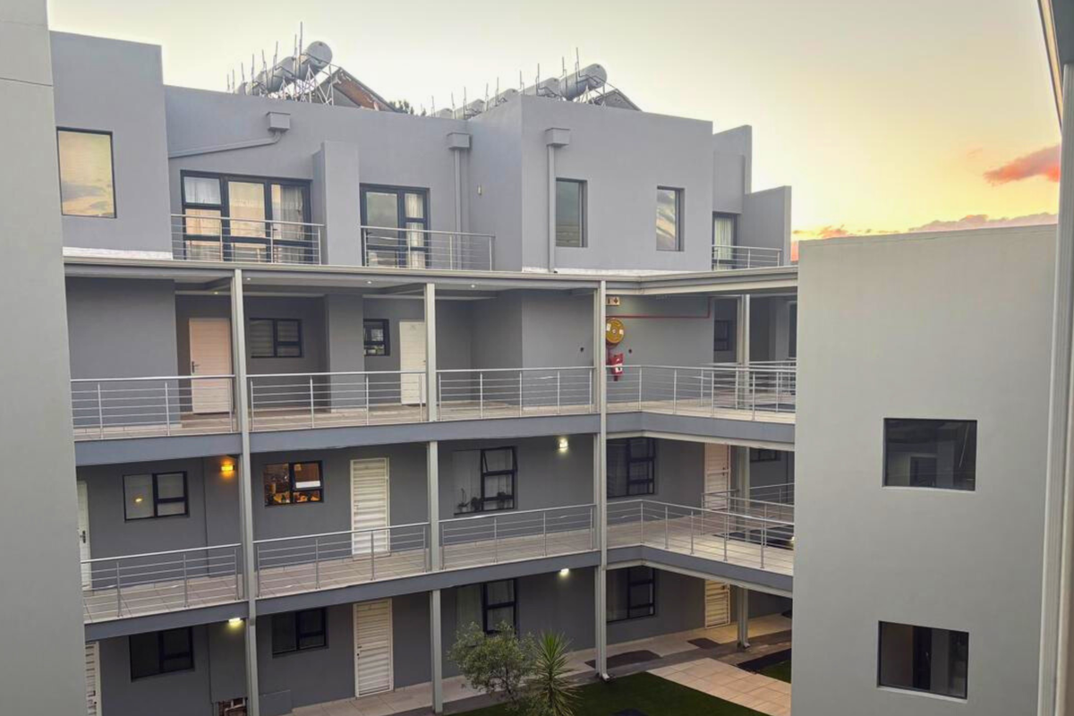 Exterior view of a modern multi-story apartment complex featuring open-air walkways, metal railings, and solar water heating systems on the roof.