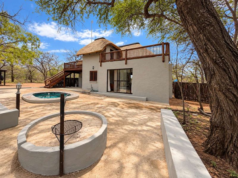Rear exterior view featuring a thatched roof dwelling, wooden balcony, plunge pool, and a built-in fire pit area set within a natural landscape.