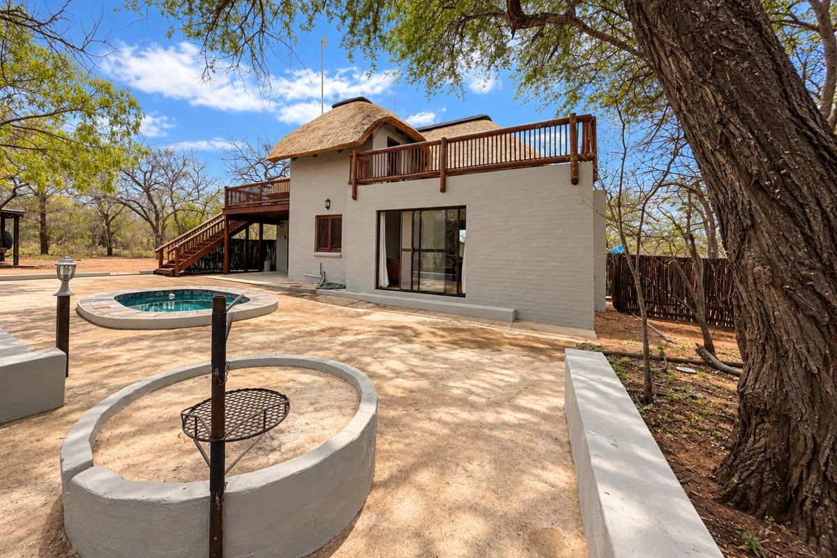 Rear exterior view featuring a thatched roof dwelling, wooden balcony, plunge pool, and a built-in fire pit area set within a natural landscape.