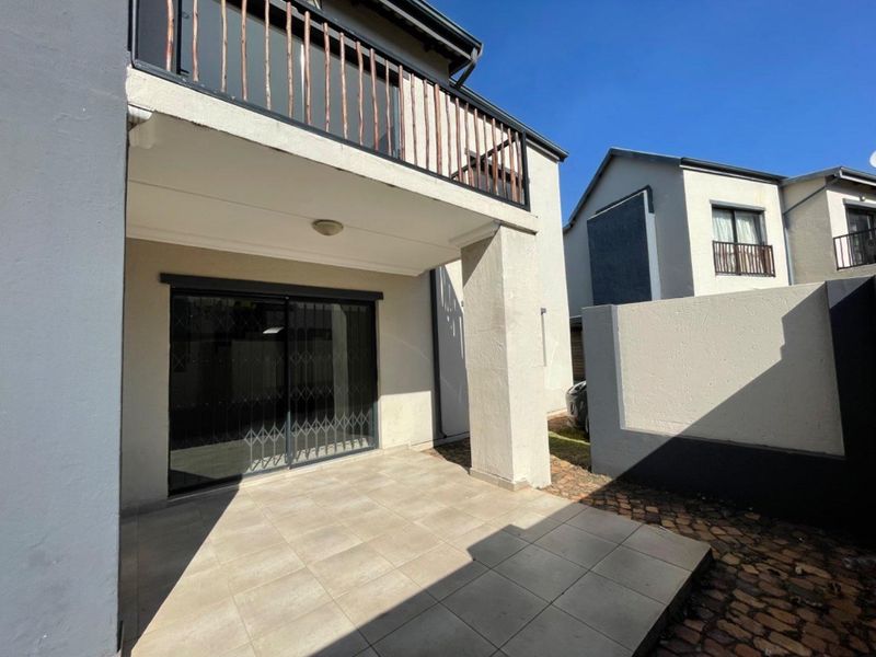 A covered ground-floor patio featuring tiled flooring and a sliding glass door with security gate, situated beneath an upper-level balcony.
