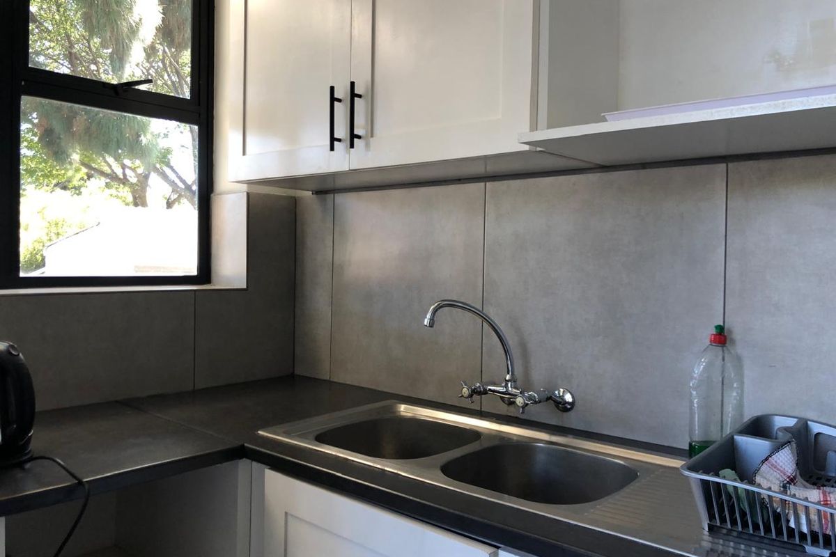 A clean, modern kitchen area featuring white cabinetry, a double bowl stainless steel sink, and grey tiled backsplash with a window for natural light.