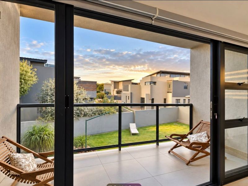 A modern covered balcony featuring tiled flooring, a sleek glass balustrade, and large sliding doors, offering a pleasant view of the surrounding residential area.