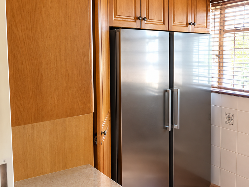 A functional kitchen area featuring warm-toned wooden cabinetry, stone-look countertops, tiled backsplash, and ceramic flooring, complemented by natural light through venetian blinds.