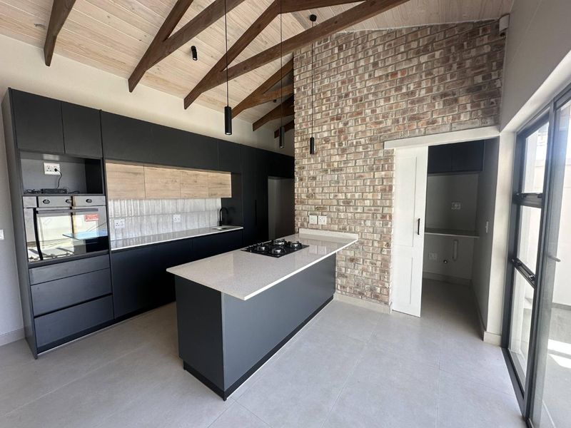 Modern kitchen featuring a central island with gas hob, sleek cabinetry, exposed brick wall, and high vaulted ceilings with timber trusses.