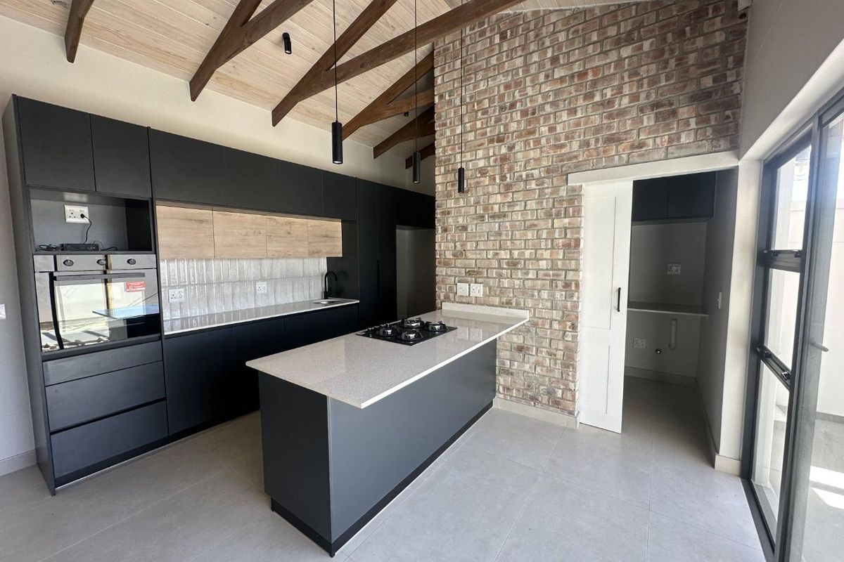 Modern kitchen featuring a central island with gas hob, sleek cabinetry, exposed brick wall, and high vaulted ceilings with timber trusses.