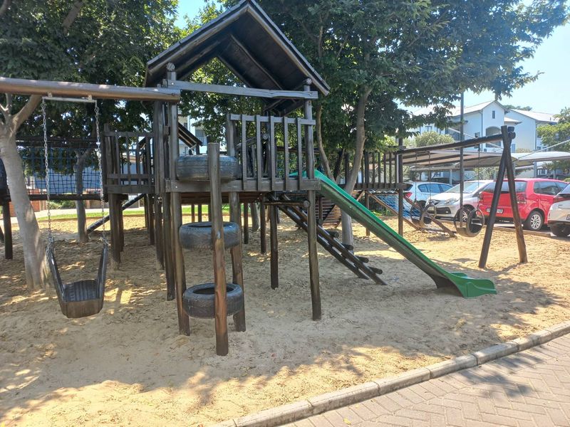 A communal outdoor playground area featuring a wooden climbing structure, slide, and swings set within a sand pit.
