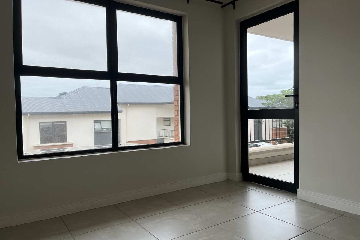 A bright, empty living area featuring large aluminium-framed windows and a glass door leading to an outdoor space, finished with neutral floor tiles.