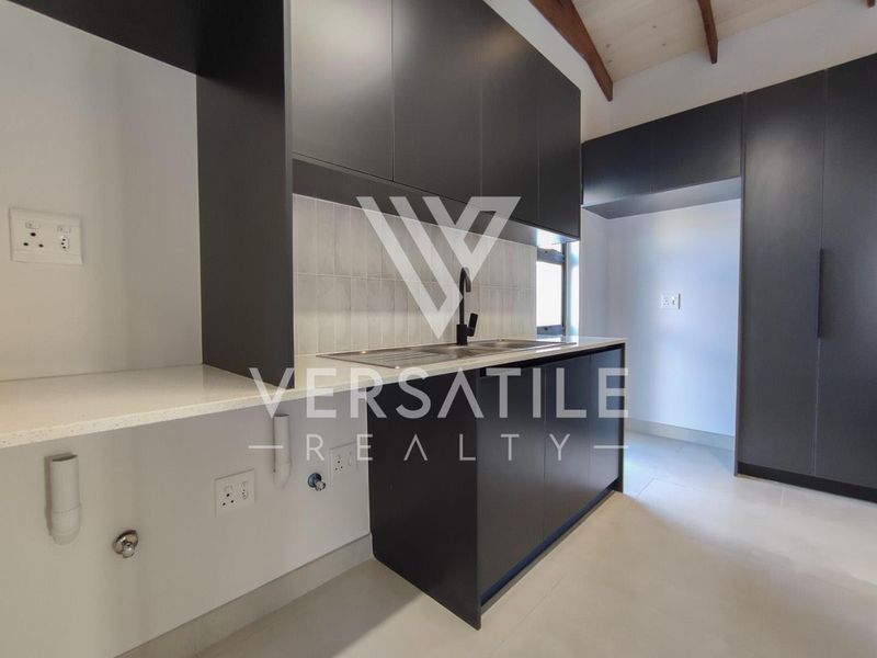 Modern kitchen featuring sleek dark cabinetry, stone countertops, a stainless steel sink, and exposed timber ceiling beams in a clean, contemporary space.