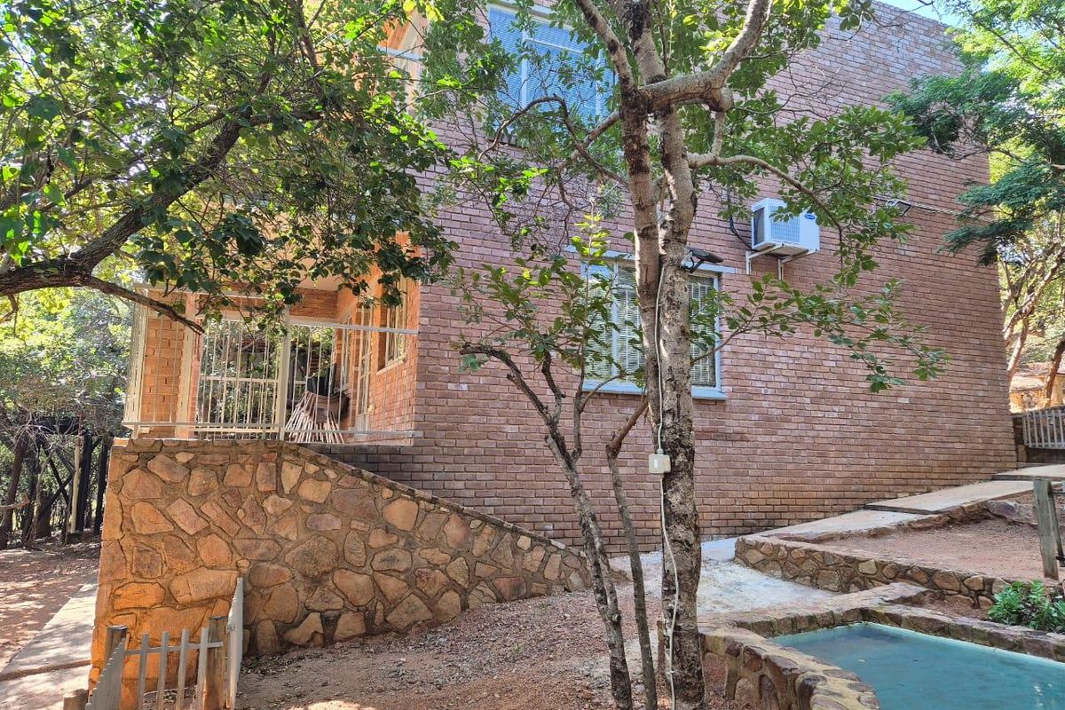 Exterior view of a multi-story brick house featuring stone retaining walls, security gates, and a small water feature in a wooded garden setting.