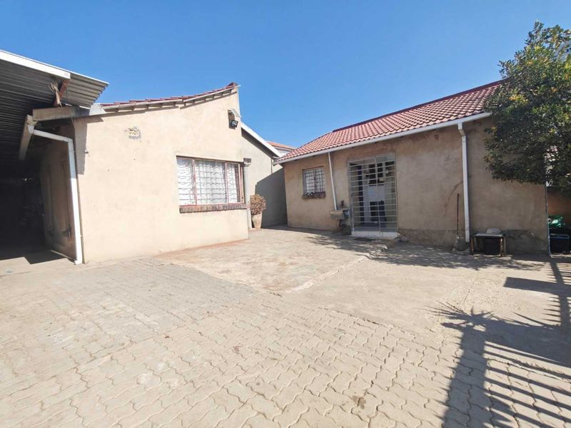 A rear exterior view of the property featuring a paved courtyard area, security bars on windows and doors, and a tiled roof structure.
