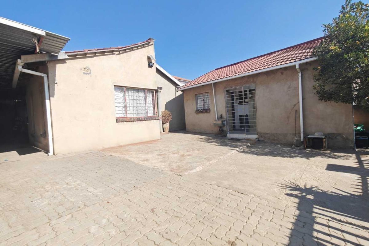 A rear exterior view of the property featuring a paved courtyard area, security bars on windows and doors, and a tiled roof structure.