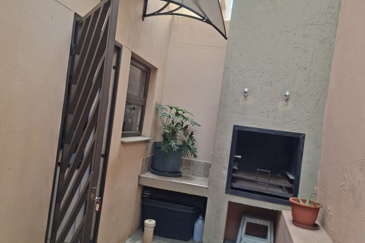 A compact rear courtyard featuring a built-in braai, paved flooring, and a protective polycarbonate awning over the entrance door.