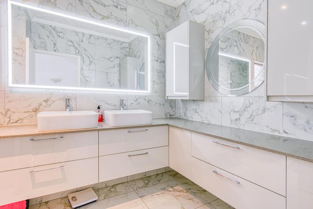 Modern bathroom featuring a double vanity with white vessel sinks, stone countertops, high-gloss cabinetry, and illuminated mirrors against marble-style wall tiling.