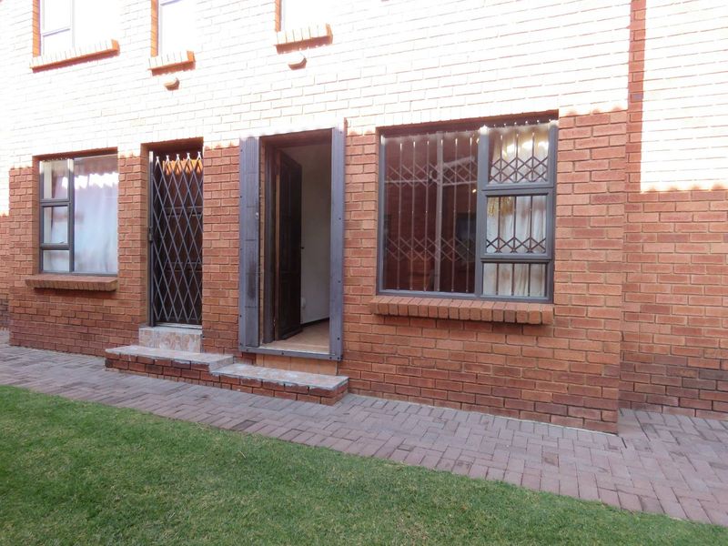 Rear exterior view of the apartment featuring a red brick facade, security gates, window security bars, and a paved walkway leading to a small lawn.