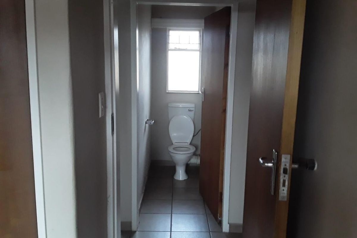 A functional bathroom featuring a standard toilet, tiled flooring, and a window providing natural light, accessed via a hallway.