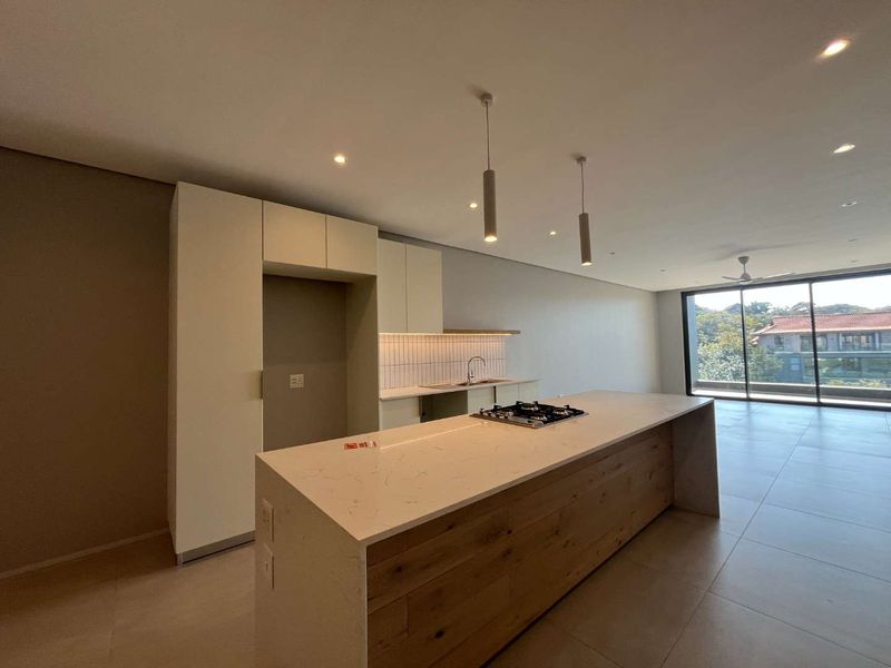 Modern open-plan kitchen featuring a large island with a gas hob, stone countertops, and sleek cabinetry, flowing into a bright living area with balcony access.