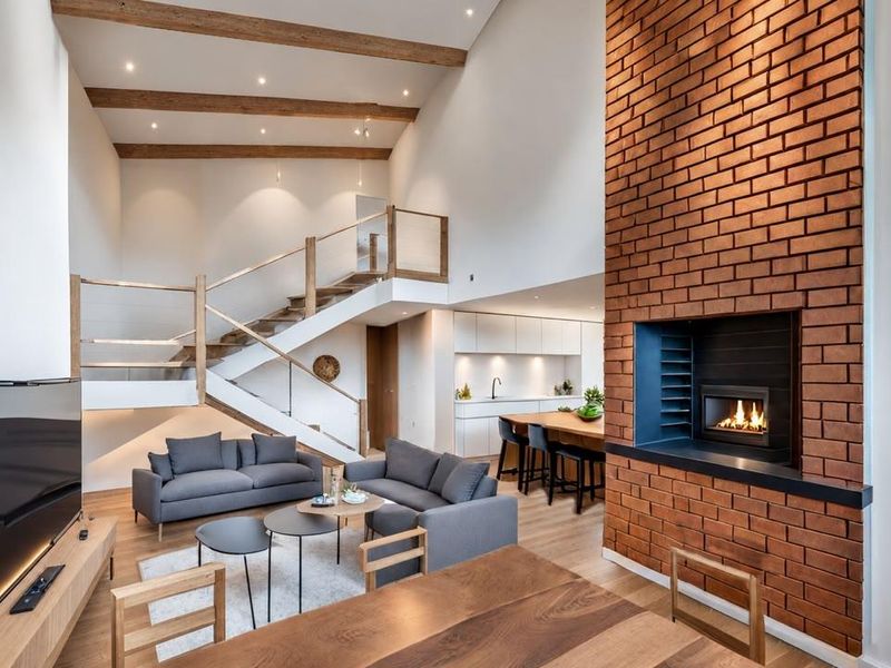 Spacious open-plan living area featuring a striking brick fireplace, exposed timber beams, a modern kitchen, and a contemporary staircase with glass balustrades.