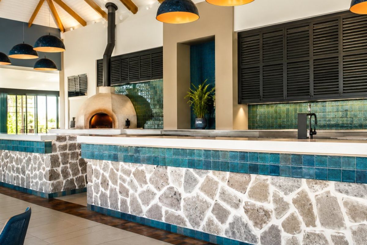 A high-end kitchen featuring a stone-clad island, integrated pizza oven, exposed timber rafters, and stylish blue pendant lighting.