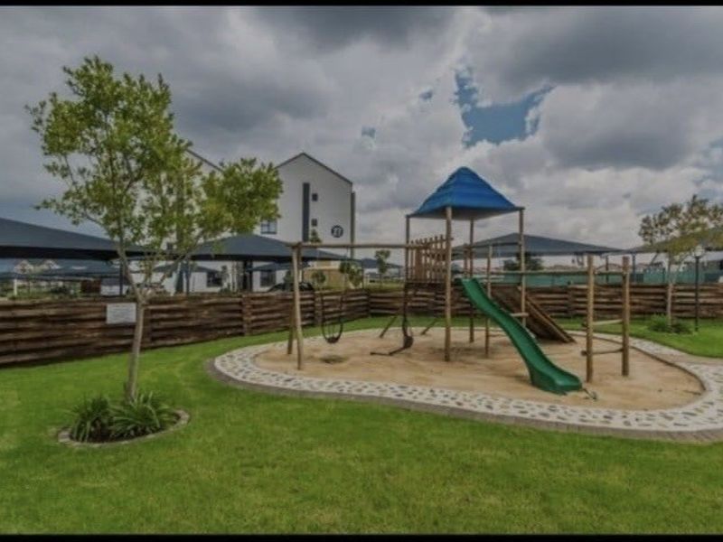A well-maintained communal garden area featuring a children's playground, manicured lawn, and timber perimeter fencing within the apartment complex.
