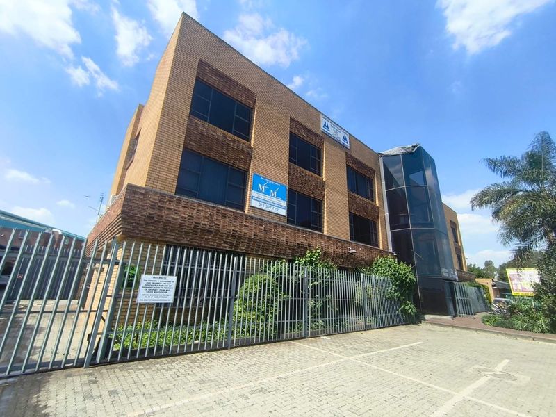 A multi-story commercial office building featuring a brick facade, glass curtain wall, and secure perimeter fencing with an adjacent paved parking area.