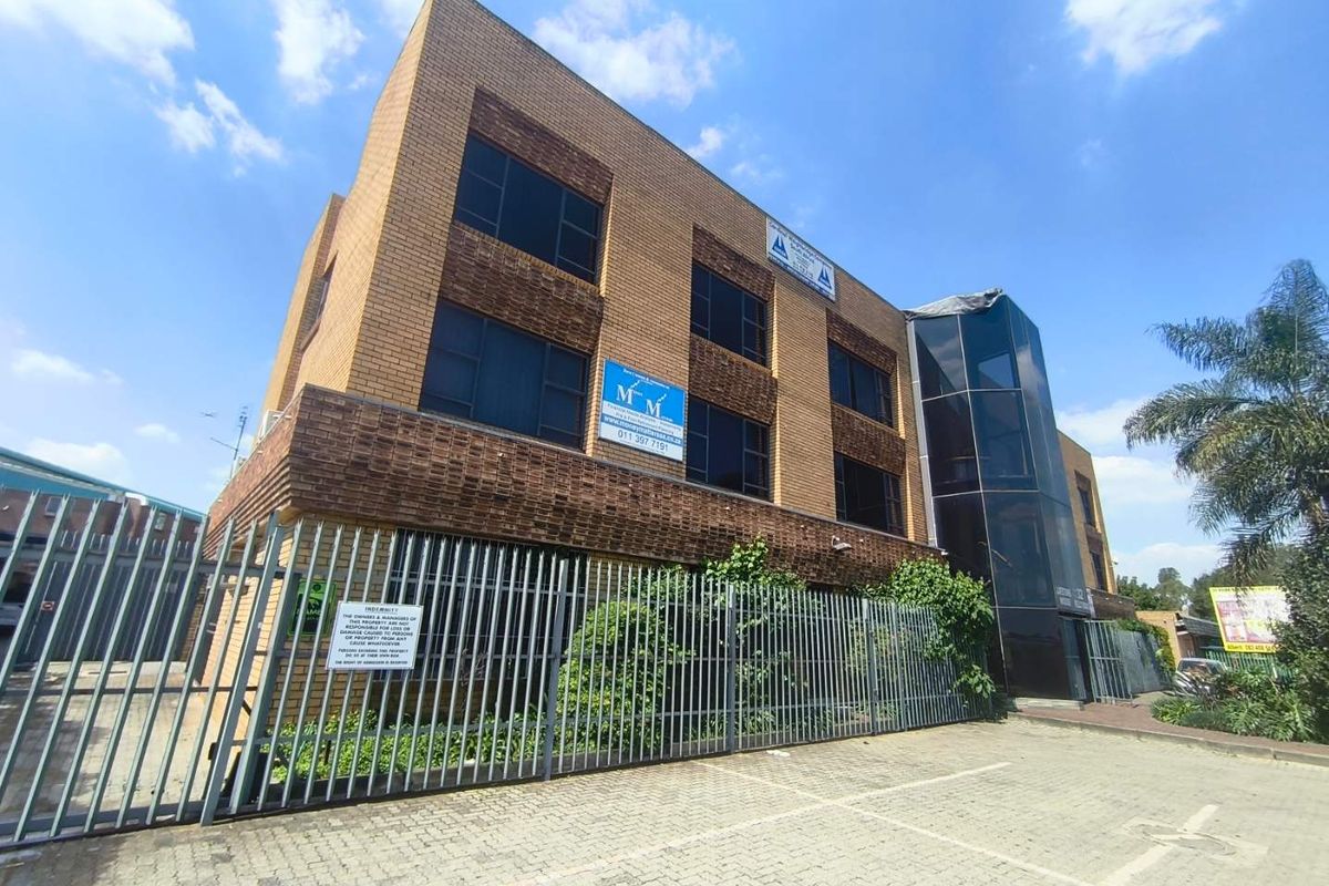 A multi-story commercial office building featuring a brick facade, glass curtain wall, and secure perimeter fencing with an adjacent paved parking area.