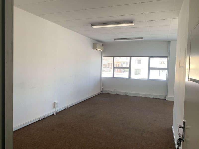 An empty, open-plan office space featuring a suspended ceiling, carpeted flooring, large windows for natural light, and a wall-mounted air conditioning unit.