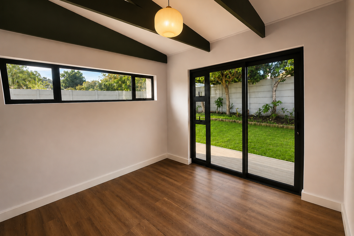 Bright, empty living area featuring exposed ceiling beams, modern laminate flooring, and large sliding glass doors providing direct access to the garden.